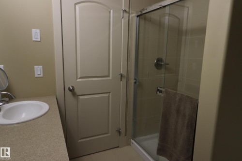 Bathroom featuring a built-in vanity with an oval sink, a light-colored countertop, a paneled interior door, and a glass-enclosed shower with a brushed metal fixture - 7492 May Common, Edmonton, AB 
