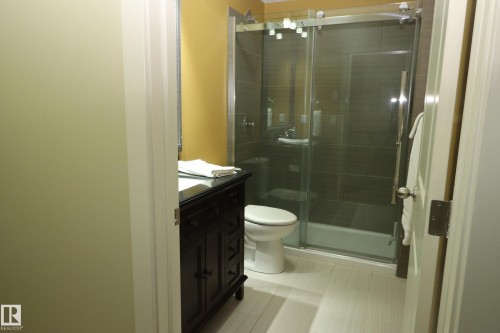 Bathroom featuring a dark wood vanity with a dark countertop, white toilet, and a glass-enclosed shower with dark tile surround - 7492 May Common, Edmonton, AB 