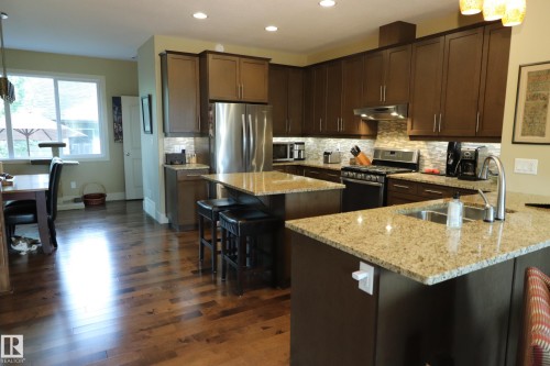 Kitchen featuring two granite-topped islands, ample dark wood-finish cabinetry, stainless steel appliances, and wood-finish flooring - 7492 May Common, Edmonton, AB 