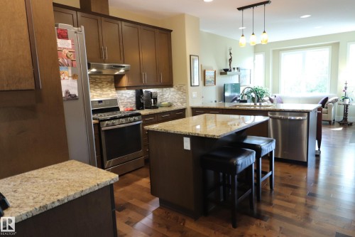Open-concept kitchen featuring dark wood cabinetry, stainless steel appliances, stone countertops, a mosaic tile backsplash, and wood-finish flooring - 7492 May Common, Edmonton, AB 