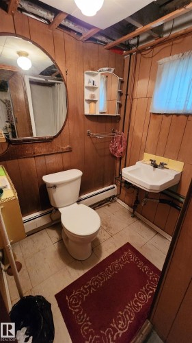Bathroom featuring wood paneling, a wall-mounted sink, a toilet, an oval mirror, and a ceiling-mounted light fixture - 9522 110 Avenue Nw, Edmonton, AB - Indoor Photo Showing Bathroom