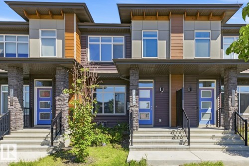 Contemporary townhome exterior featuring stone pillars, a vibrant blue entry door with frosted glass panels, black metal railings, and a multi-textured facade with wood-look and gray paneling accents - 1364 Keswick Drive Sw, Edmonton, AB 