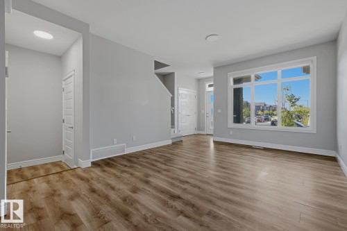 Open-concept living area featuring wood-finish flooring, a large multi-pane window, light gray wall paint, white trim, and a white interior door - 1364 Keswick Drive Sw, Edmonton, AB 