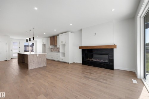 Expansive main level featuring wood-finish flooring, a contemporary fireplace with a black tile surround and wood mantel, a kitchen island with pendant lighting, and abundant natural light from large windows - 39 Elsinore Place, Edmonton, AB 