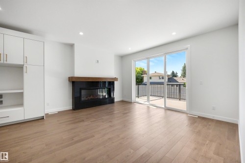 Living area featuring wood-finish flooring, a modern black fireplace with a wood mantle, built-in white cabinetry with gold hardware, recessed lighting, and a sliding glass door opening to an outdoor balcony - 39 Elsinore Place, Edmonton, AB 