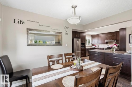 Open-concept dining area with wood-finish flooring, featuring a contemporary ceiling light fixture and a spacious kitchen with dark wood cabinetry, stainless steel appliances, and light-colored countertops - 4007 45 Street, Stony Plain, AB 