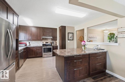 Kitchen featuring dark wood cabinetry, stainless steel appliances, a tile backsplash, and a central island with a stone-finish countertop - 4007 45 Street, Stony Plain, AB 