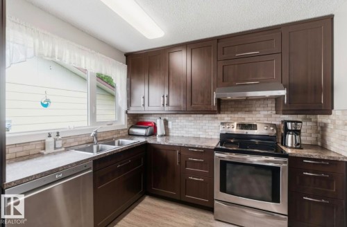 Kitchen featuring rich wood cabinetry, stainless steel appliances, a double basin sink, stone-look countertops, and a tiled backsplash - 4007 45 Street, Stony Plain, AB 