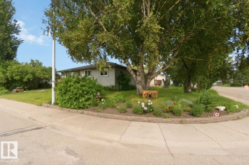Corner lot featuring a mature tree, landscaped flower beds with retaining wall, a manicured lawn, and a light-colored exterior residence - 4007 45 Street, Stony Plain, AB 