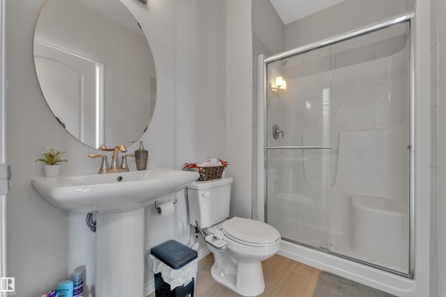 Full bathroom featuring a white pedestal sink with brass-finish faucet, a circular wall mirror, wood-finish flooring, and a shower with sliding glass doors - 2246 54 Street, Edmonton, AB 