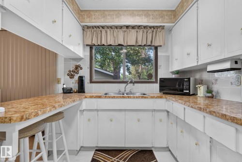 Kitchen featuring L-shaped countertop, white cabinetry with gold-tone hardware, a double-basin sink, and a tiled backsplash - 4712 131 Avenue Nw, Edmonton, AB 