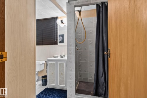 Bathroom featuring a tiled shower stall with a dark shower curtain, a light wood vanity with a white countertop, and a window with dark blinds - 4712 131 Avenue Nw, Edmonton, AB 