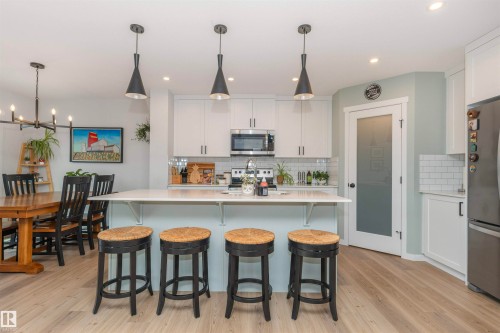 Open-concept kitchen featuring an island with counter seating, stainless steel appliances, white subway tile backsplash, and light wood-finish flooring - 102 Horton Way, Ardrossan, AB 