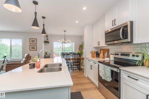 Open-concept kitchen featuring a large island with a double sink, wood-finish flooring, white cabinetry, stainless steel appliances, and a subway tile backsplash - 102 Horton Way, Ardrossan, AB 