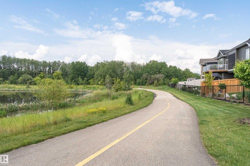 Paved pathway alongside a naturalized pond, bordered by manicured lawn and mature trees, with modern homes featuring balconies and privacy fencing visible in the background - 102 Horton Way, Ardrossan, AB 
