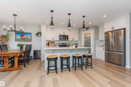 Open-concept kitchen featuring an island with seating, stainless steel appliances, white cabinetry, subway tile backsplash, and light wood-finish flooring - 102 Horton Way, Ardrossan, AB 