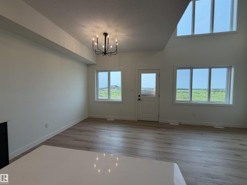 Spacious living area featuring light wood-finish flooring, a contemporary black chandelier, multiple large windows, a white paneled door, and a high vaulted ceiling with additional upper windows - 115 Pierwyck Loop, Spruce Grove, AB 
