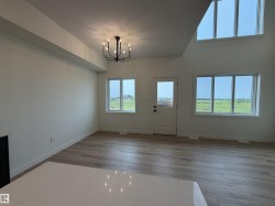 Spacious living area featuring light wood-finish flooring, a contemporary black chandelier, multiple large windows, a white paneled door, and a high vaulted ceiling with additional upper windows - 