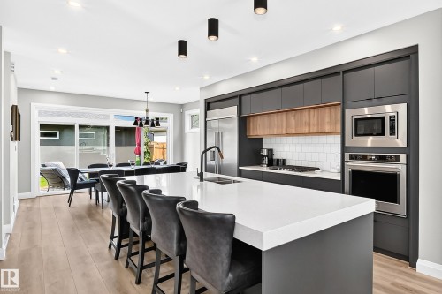 Modern kitchen featuring a large white countertop island with integrated seating, dark flat-panel cabinetry, stainless steel appliances, and wood-finish flooring - 12631 52 Avenue, Edmonton, AB 
