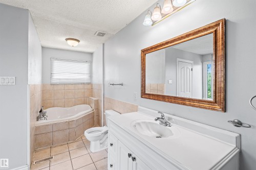 Bathroom featuring a built-in corner bathtub with tiled surround, a white vanity with an integrated sink, a large framed mirror, and a window with blinds - 11507 141 Avenue Nw, Edmonton, AB - Indoor Photo Showing Bathroom