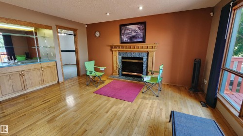 Spacious common area featuring wood-finish flooring, a fireplace with a wood mantle and dark tile surround, a built-in wet bar with mirrored backing and glass shelving, and large windows providing abundant natural light - 2708 44A Avenue, Edmonton, AB 