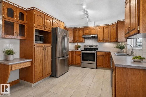 Kitchen featuring wood cabinetry, stainless steel appliances, a built-in desk area, a double basin sink, and light-toned tile flooring - 5612 Mcleod Road, Edmonton, AB 