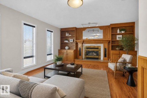 Living area featuring wood-finish flooring, a central fireplace with a decorative mantel and surround, built-in wooden shelving, and two tall windows with blinds - 5612 Mcleod Road, Edmonton, AB 