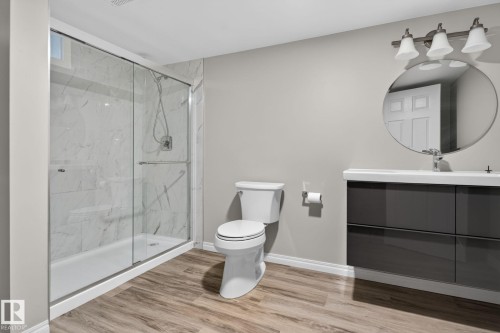 Contemporary bathroom featuring a spacious glass-enclosed shower with white marble-look tile, a floating vanity with a white countertop and dark gray cabinetry, a round wall-mounted mirror, and wood-finish flooring - 5612 Mcleod Road, Edmonton, AB 