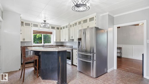 Kitchen featuring a central island with a light-toned countertop, white cabinetry with clear-fronted upper doors, stainless steel appliances, and tile flooring - 10115 104 Street, Westlock, AB 