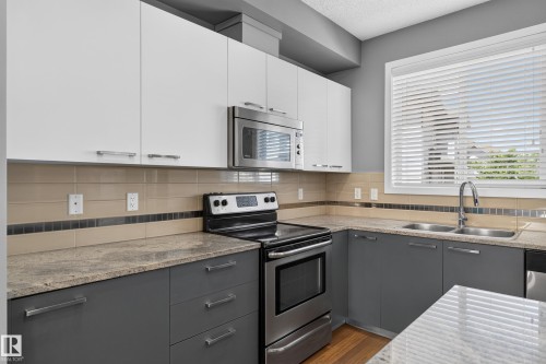 Modern kitchen featuring two-tone cabinetry, stainless steel appliances, speckled countertops, a tile backsplash with decorative accent, and wood-finish flooring - 24 655 Watt Boulevard, Edmonton, AB 