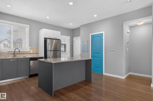 Modern kitchen featuring a central island with a light-toned countertop, stainless steel appliances, dark gray cabinetry, and wood-finish flooring - 24 655 Watt Boulevard, Edmonton, AB 