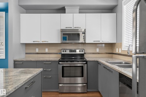 Kitchen featuring a stainless steel appliance suite, two-tone cabinetry, stone countertops, a tiled backsplash with accent stripe, and wood-finish flooring - 24 655 Watt Boulevard, Edmonton, AB 