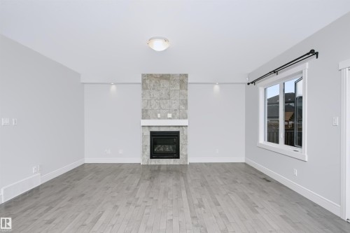 Living area featuring a floor-to-ceiling tiled fireplace with a white mantel, wood-finish flooring, and a large window with black hardware - 1235 164 Street, Edmonton, AB 