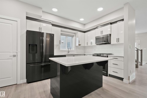 Modern kitchen featuring a central island with a white countertop and black base, white upper and lower cabinetry, a black appliance suite, and light wood-finish flooring - 383 Charlesworth Drive, Edmonton, AB 