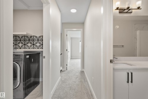 Hallway with plush carpet flooring, a dedicated laundry area featuring decorative tile backsplash and a floating shelf, and a bathroom entrance revealing a vanity with a white countertop and dark hardware - 383 Charlesworth Drive, Edmonton, AB 