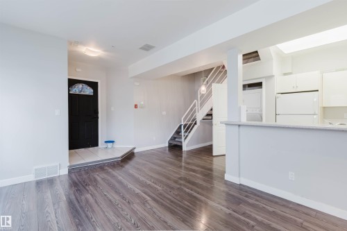 Open-concept living area featuring wood-finish flooring, a contemporary staircase with white railings, and a kitchen with white cabinetry and appliances - 202 10939 82 Avenue, Edmonton, AB 