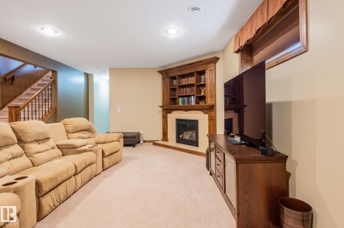 Carpeted recreation room featuring a corner fireplace with a tile surround and wood mantel, built-in shelving, a wood-finish media console, and a staircase with wood newel posts and handrails - 19 Newton Place, St. Albert, AB - Indoor With Fireplace