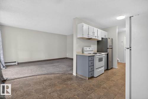 Kitchen area featuring tile flooring, white upper cabinetry, blue lower cabinetry, a white electric range, and a stainless steel refrigerator - 106 237 Woodvale Road W, Edmonton, AB 