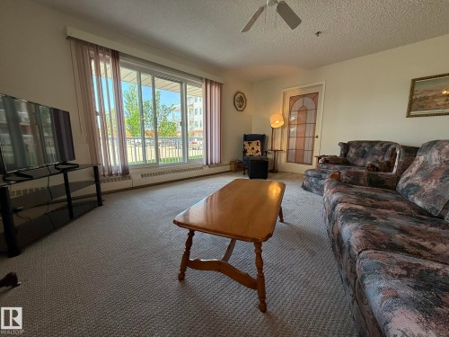 Living area featuring a textured ceiling with a ceiling fan, light-colored carpeting, and a large window with sheer curtains - 102 10903 21 Avenue, Edmonton, AB - Indoor Photo Showing Living Room