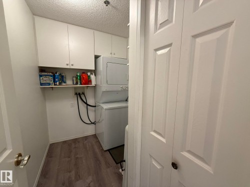 Dedicated laundry area featuring a stacked washer and dryer, wood-finish flooring, white overhead cabinetry, and a wall-mounted shelf - 102 10903 21 Avenue, Edmonton, AB - Indoor