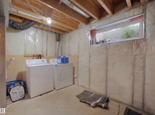 Unfinished basement space featuring exposed wooden ceiling joists, a dedicated laundry area with hookups, and a horizontal window with a corrugated well - 8111 7 Avenue Sw, Edmonton, AB 