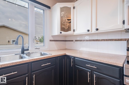 Kitchen featuring a double stainless steel sink, a gooseneck faucet, two-tone cabinetry with black base cabinets and white upper cabinets, a light-toned countertop, and a white tile backsplash with a decorative mosaic accent stripe - 11415 142 Avenue, Edmonton, AB 