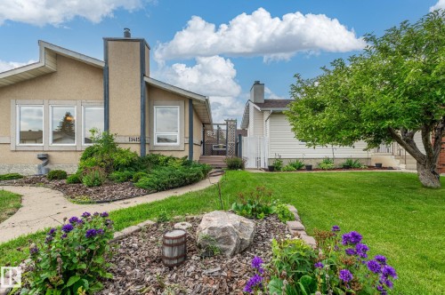 Exterior featuring tan stucco siding, an angled roofline, multiple windows, a prominent chimney, and a landscaped front yard with a manicured lawn and mature trees - 11415 142 Avenue, Edmonton, AB 