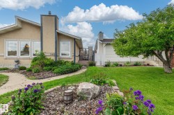 Exterior featuring tan stucco siding, an angled roofline, multiple windows, a prominent chimney, and a landscaped front yard with a manicured lawn and mature trees - 