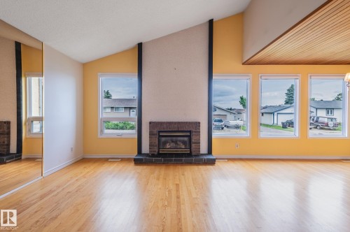 Spacious interior featuring a brick fireplace with a dark hearth, wood-finish flooring, a vaulted ceiling with wood paneling, and multiple windows - 11415 142 Avenue, Edmonton, AB 