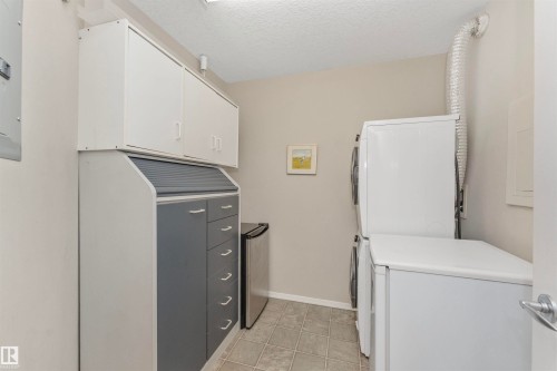Functional utility area featuring a stacked washer and dryer, tile flooring, and ample built-in cabinetry with both white and gray finishes - 130 9008 99 Avenue Nw, Edmonton, AB 