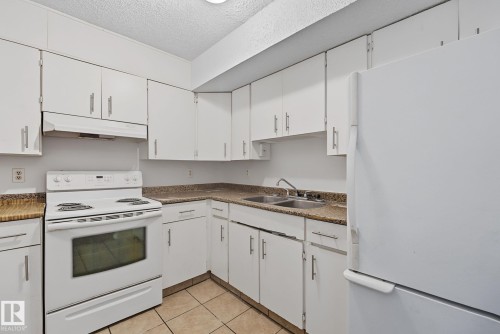 White kitchen featuring extensive cabinetry, L-shaped countertops, double basin sink, and tile flooring - #6E 11608 28 Av Nw Nw, Edmonton, AB - Indoor Photo Showing Kitchen With Double Sink