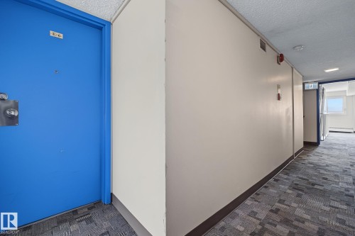 Corridor entrance featuring a vibrant blue door with chrome hardware, textured acoustic ceiling, light-colored walls, and patterned carpet flooring - #6E 11608 28 Av Nw Nw, Edmonton, AB - Indoor Photo Showing Other Room