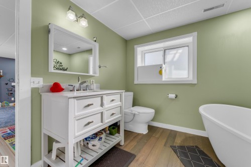 Bathroom featuring a white vanity with a rectangular sink, light-colored walls, wood-finish flooring, a white freestanding bathtub, and a window - 1316 35 Street, Edmonton, AB 