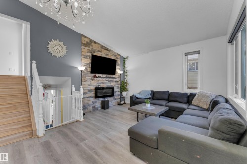 Contemporary living space featuring a stacked stone fireplace surround with an integrated electric fireplace, light wood-finish flooring, a crystal chandelier, painted wood stair railings, and an accent wall - 1316 35 Street, Edmonton, AB 